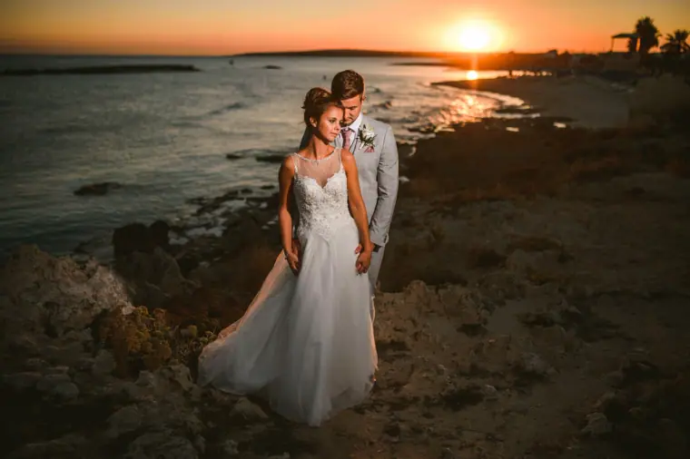 Naomi and Dale standing on the rocky shore as the sun sets over the sea, Ayia Napa