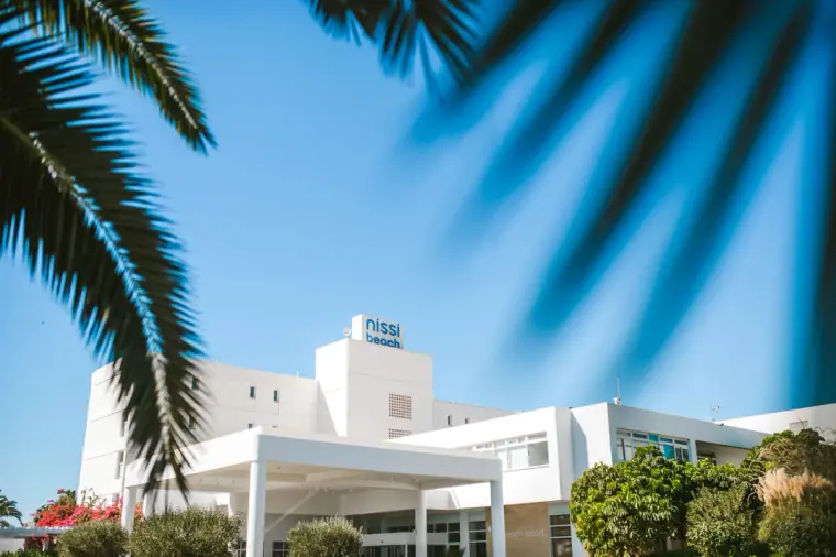 White Nissi Beach Resort building under blue sky, framed by palm fronds, Ayia Napa