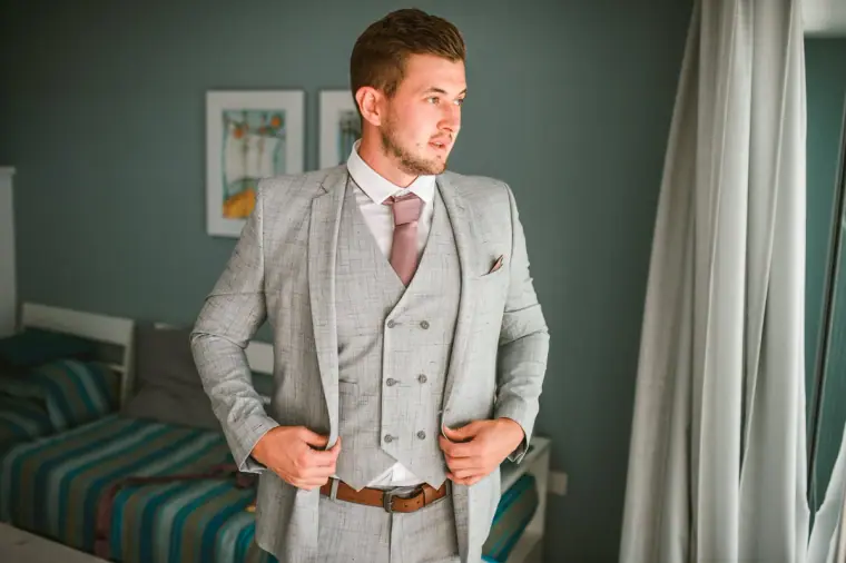 Groom in a light grey three-piece suit buttoning his jacket in the hotel room
