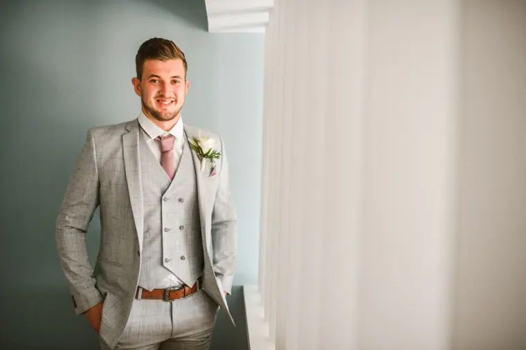 Groom in a grey three-piece suit with a pink tie standing by the window