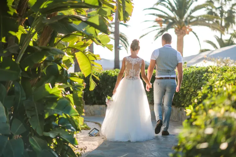 Bride and groom walking away hand in hand along a palm-lined garden path