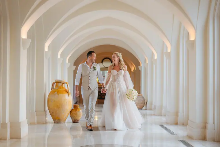 Jade and Peter walking hand in hand through the white arched colonnade at Anassa