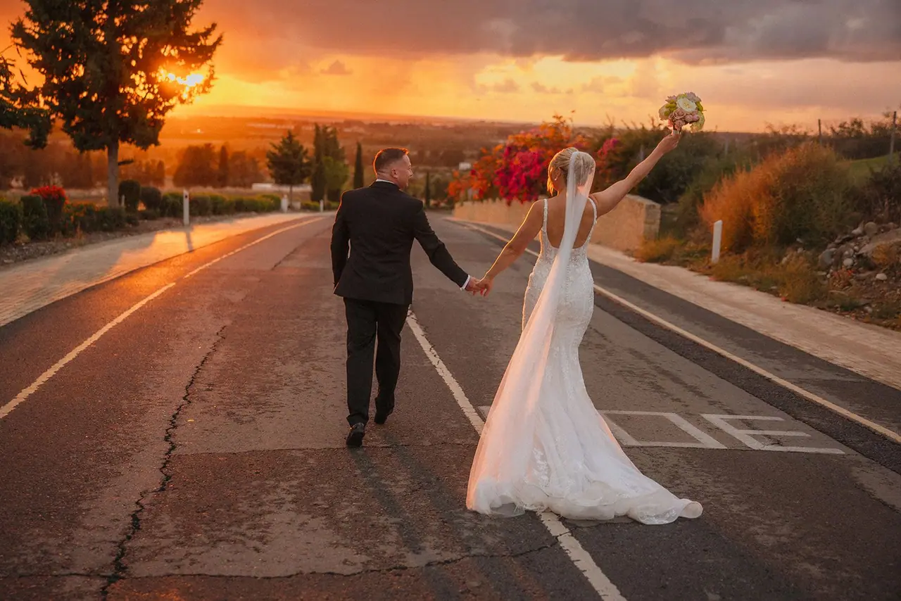 Zoe raising her bouquet as she and Oliver walk down the Kouklia road into the sunset after their Liopetro wedding