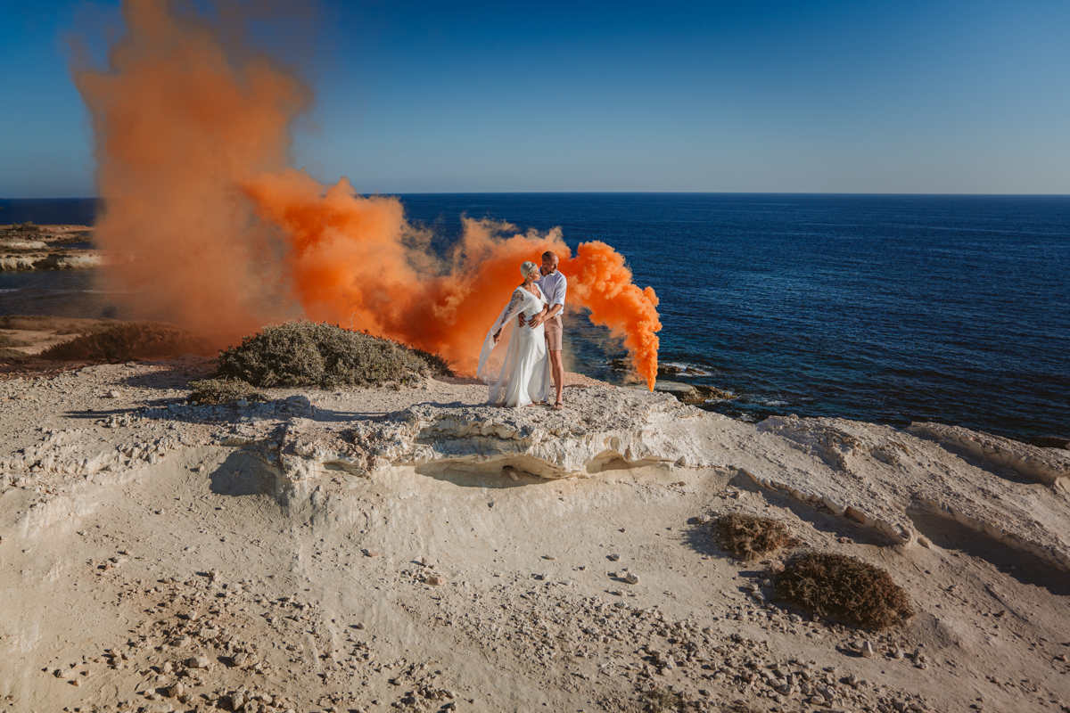 An orange flare contrasts the blue skies and sea - a dramatic backdrop for the newlyweds who embrace in front of it